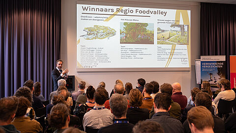 Deelnemers in de zaal kijken naar de presentatie van Hugo de Jonge. Hij toont de dia waarop de drie ontwerpen staan van de drie ontwerpteams.  