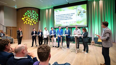 Politci staan voor in de zaal te debateren. 