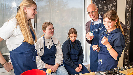 Wethouder 't Jong staat in de keuken met de leerlingen. Ze zijn aan het koken.
