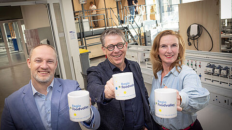 Dennis Gudden, Joop Hazeleger en Diane Moerkerk houden de koffiemok vast. Zij staan in een werkplaats. 