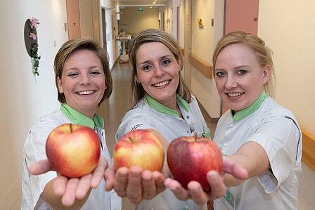 Op de foto staan drie verpleegsters met een appel in de hand.