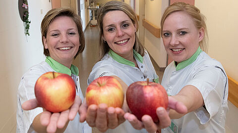 Op de foto staan drie verpleegsters met een appel in de hand.
