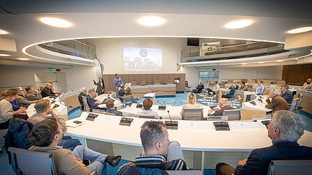 De deelnemers zitten in de raadszaal Ede. Een overzichtsfoto van de zaal. 