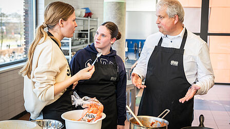 Tijd om te proeven. De wethouder en twee leerlingen proeven het gerecht in de keuken.