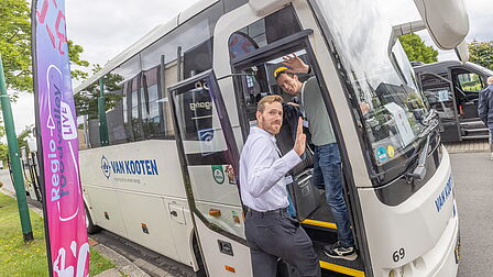 Deelnemers stappen in de bus vanaf de Basiliek in Veenendaal. Man zwaait naar de camera. 
