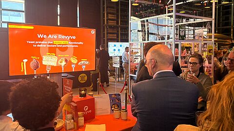 De eb-leden en gasten staan bij de stand van Revyve. Uitleg wordt gegeven en er wordt geproefd.