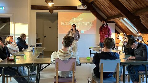 Kick-off bijeenkomst van de boardroom is gestart. De leerlingen zitten in de zaal en luisteren naar het welkomstwoord van trainer Demi. 